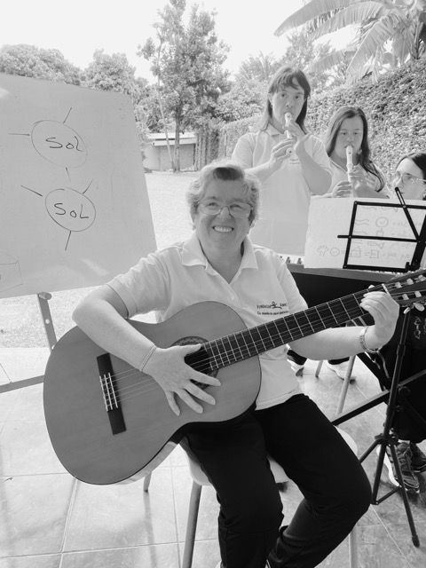 Una docente sonriente toca la guitarra mientras dos estudiantes participan